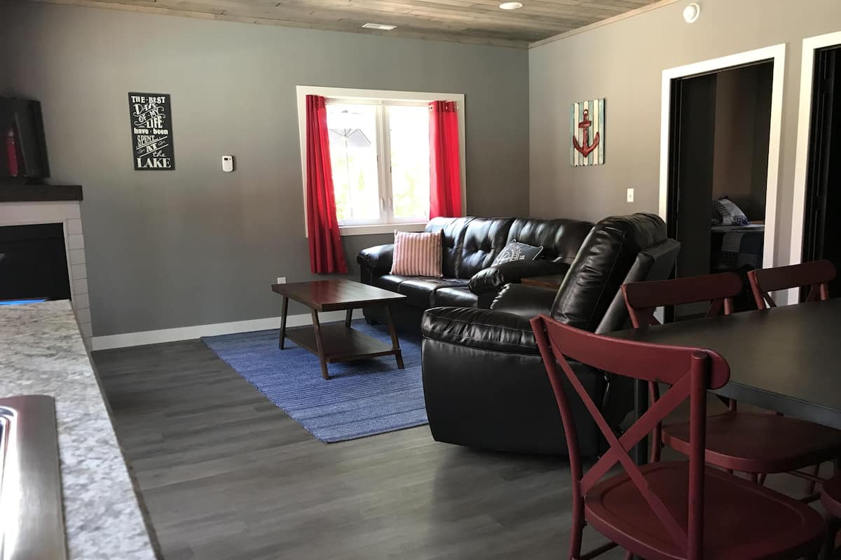 A living room with a black leather sofa, a wooden coffee table, and a blue rug. There is a dining table with red chairs. The room has gray walls and a window with red curtains.