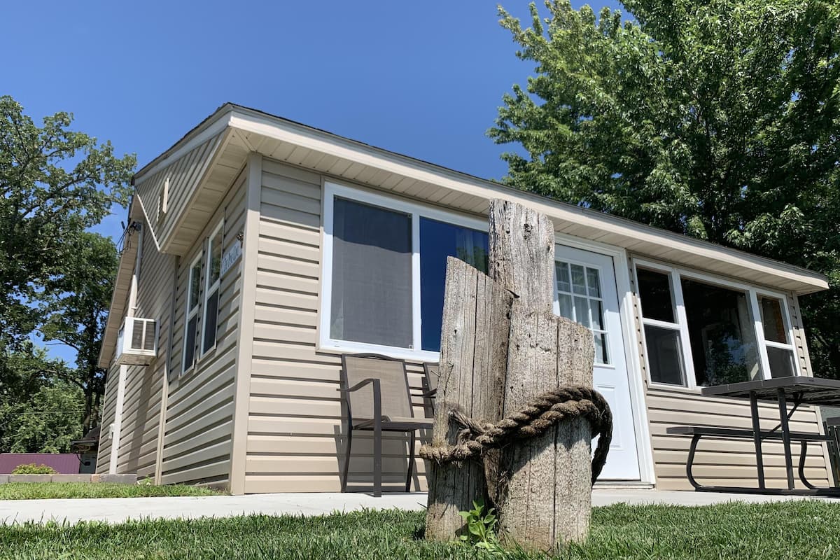 A single-story house with a beige exterior, a large window, and a tree stump in the front yard.