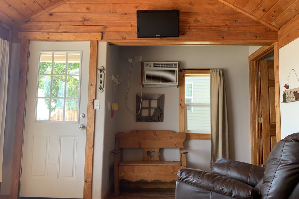 A cozy cabin interior with a wooden ceiling, a television mounted above a bench, and a leather sofa.