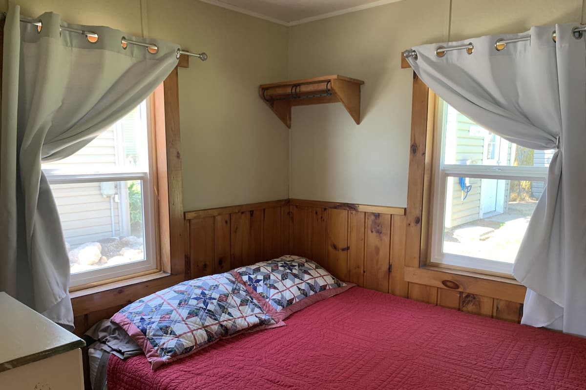 A bedroom with a wooden headboard, a red quilted bedspread, and a patterned pillow. There are two windows with curtains, and a wooden shelf above the bed.