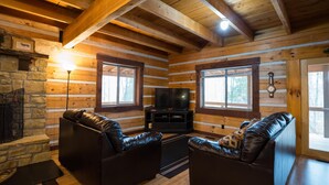 Ein Holzblockhaus-Interieur mit Kamin aus Stein, Ledersofas, einem Fernseher und großen Fenstern.