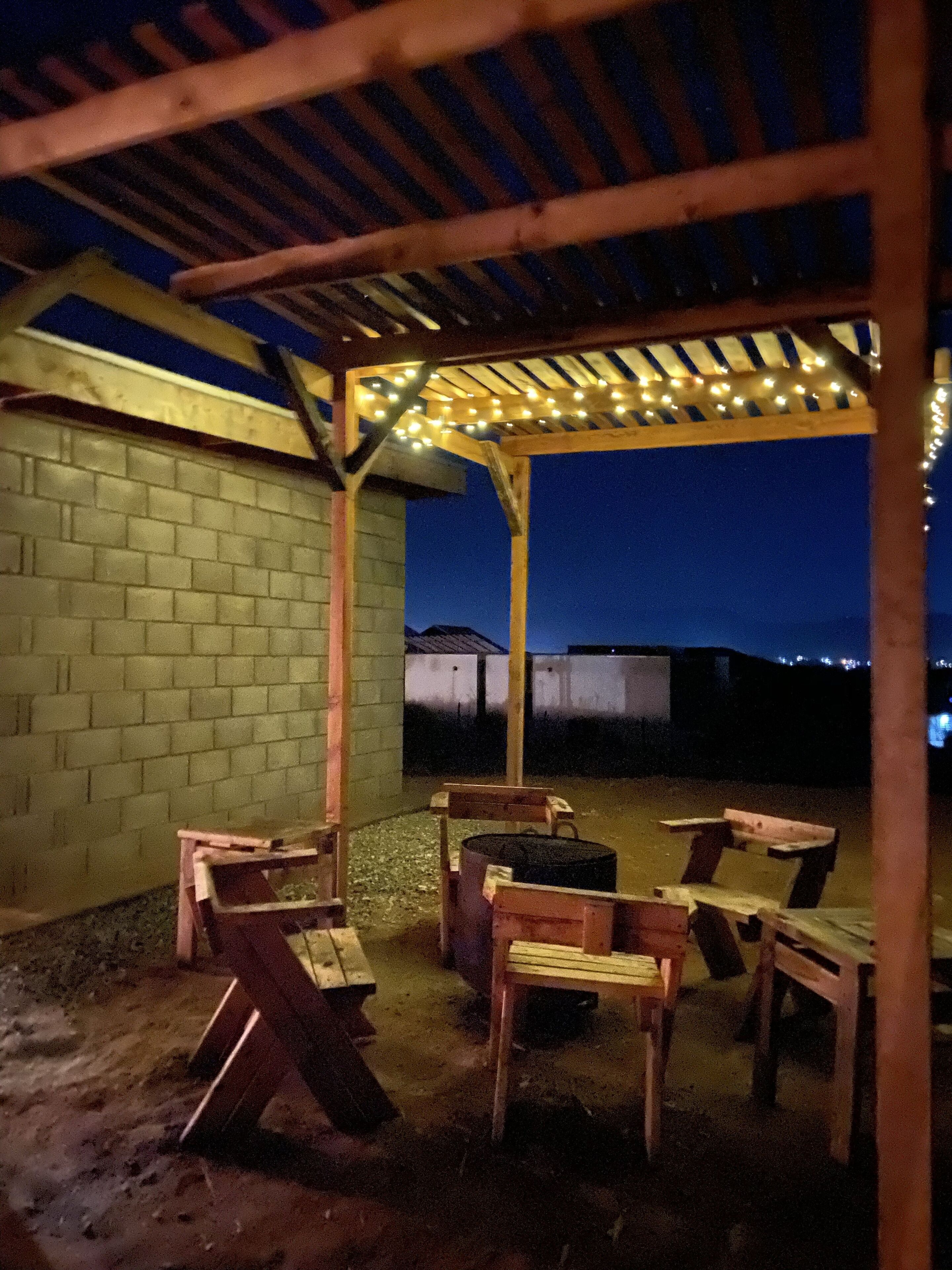 Un patio exterior con bancos de madera y una pérgola adornada con luces colgantes.