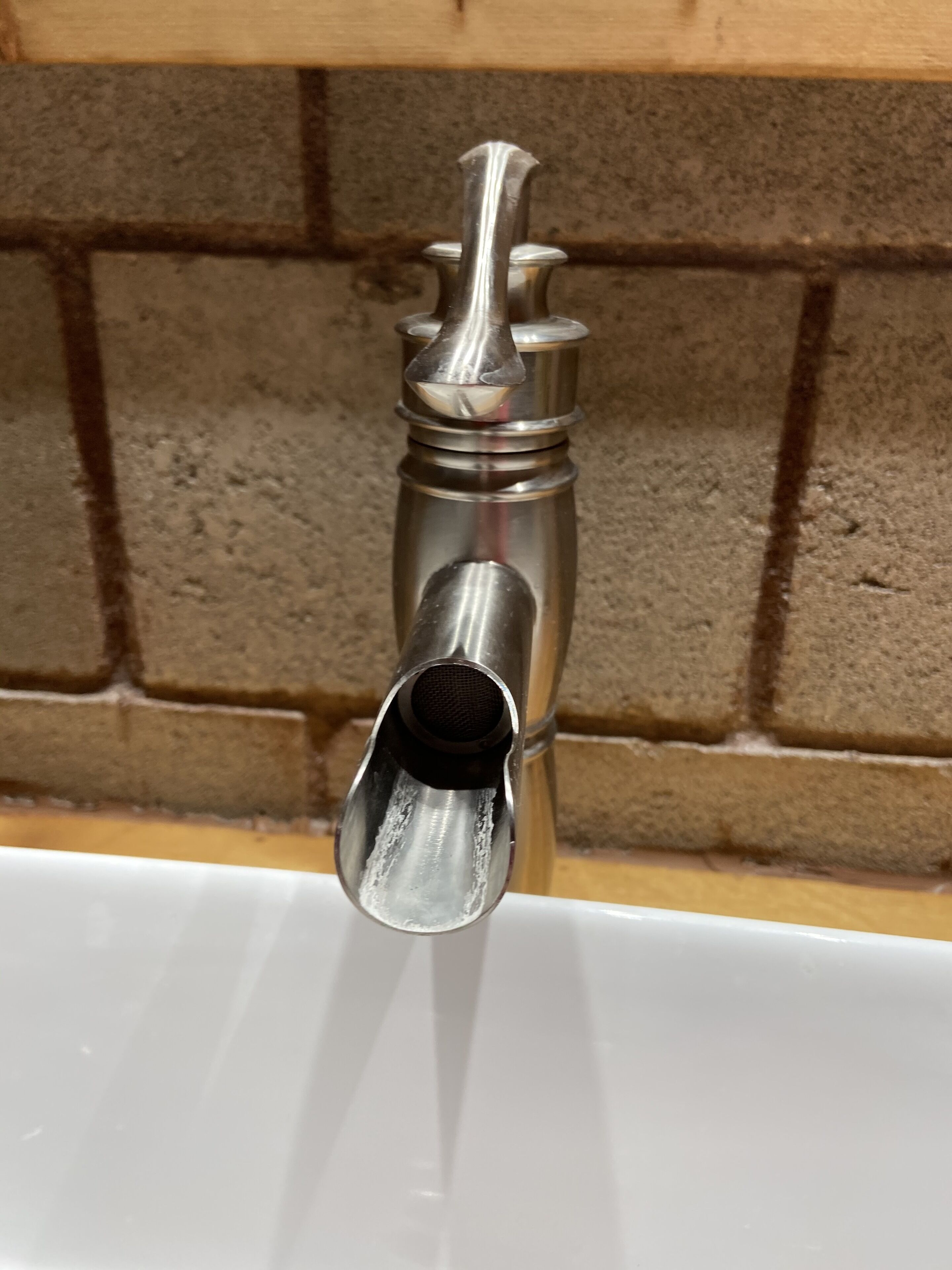 Een metalen kraan met één hendel en een tuit, gemonteerd op een witte wasbak voor een bakstenen muur.
