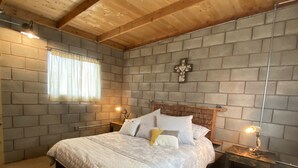 A bedroom with a wooden ceiling, a brick wall, a bed with a white quilt, two bedside tables with lamps, and a patterned rug. - Sn Tony Hotel & Spa (Valle de Guadalupe)
