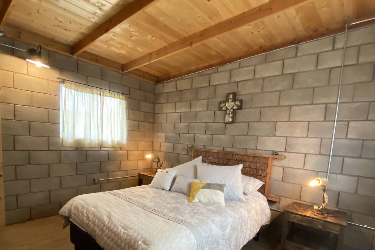 A bedroom with a wooden ceiling, a brick wall, a bed with a white quilt, two bedside tables with lamps, and a patterned rug.