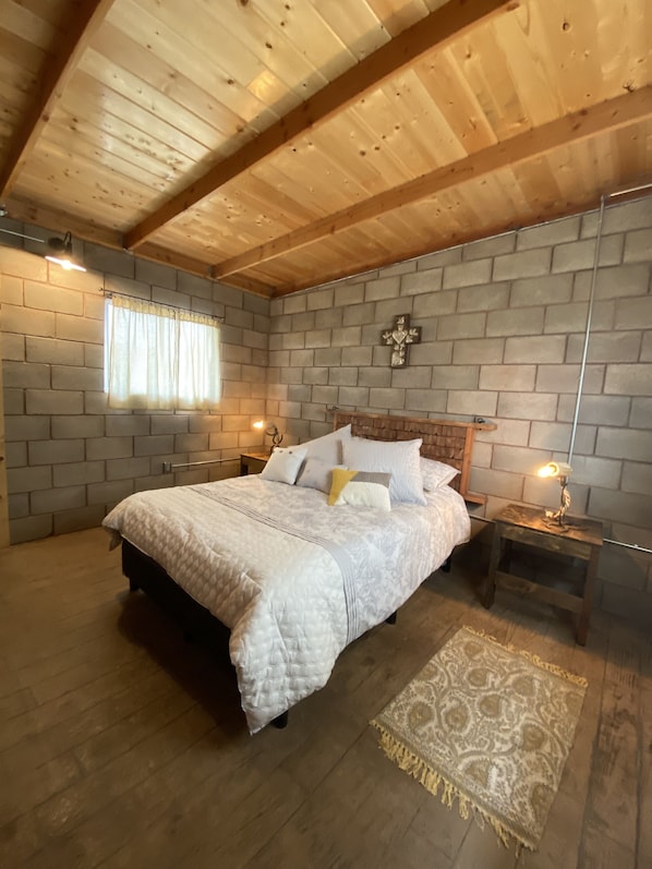 A bedroom with a wooden ceiling, a brick wall, a bed with a white quilt, two bedside tables with lamps, and a patterned rug. - Sn Tony Hotel & Spa (Valle de Guadalupe)