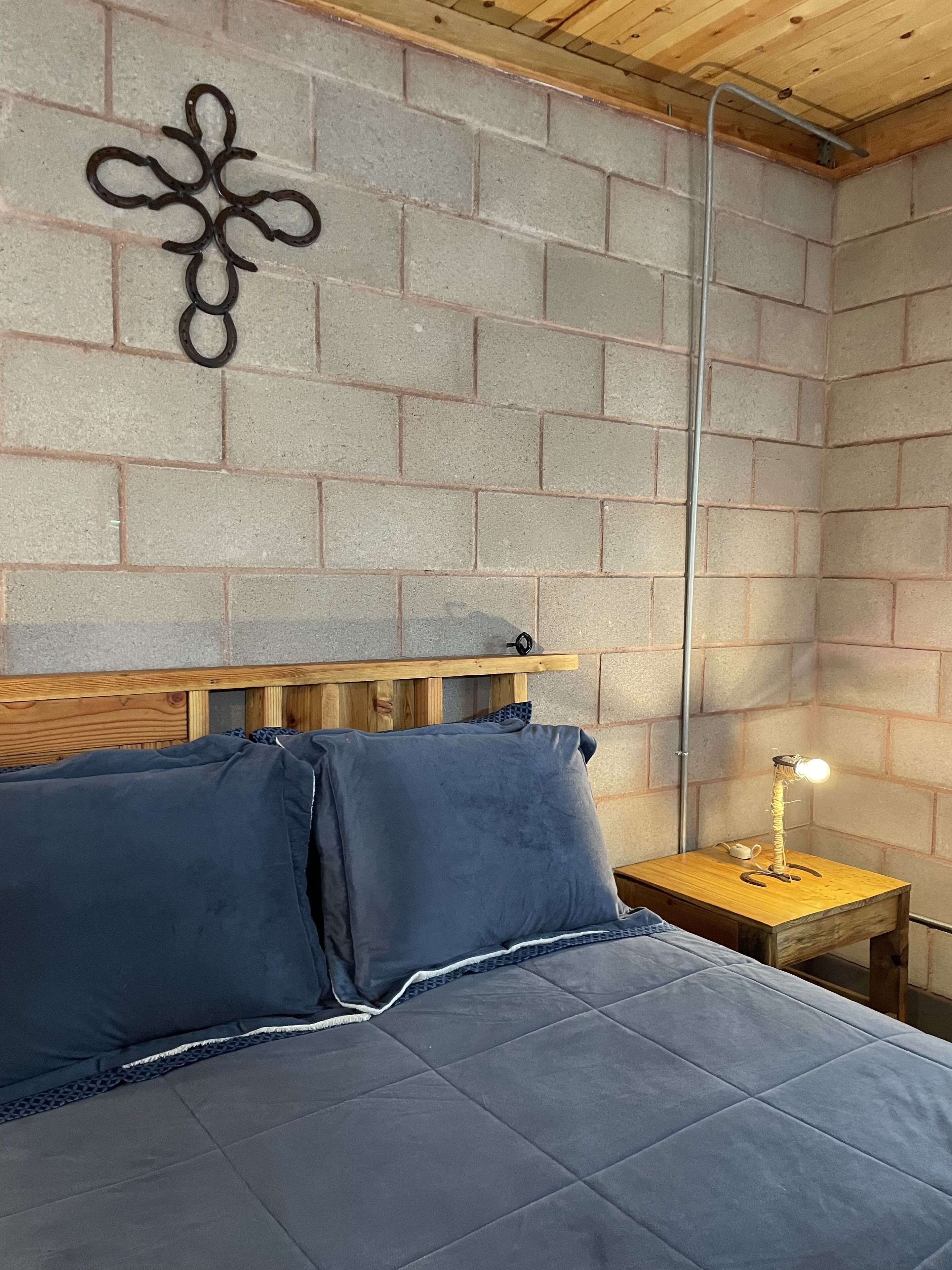 Un dormitorio con pared de ladrillo, cabecera de madera, cama con ropa de cama azul y mesita de noche con lámpara.