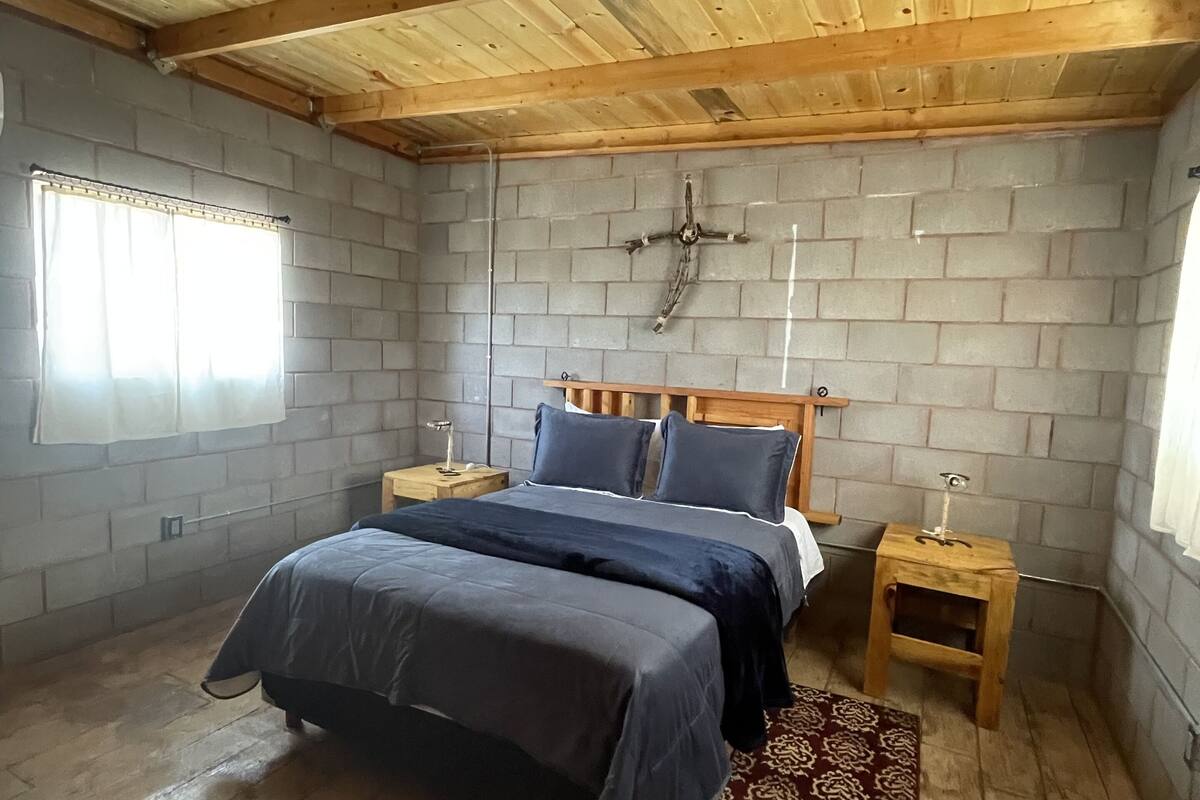 A bedroom with a wooden ceiling, a brick wall, a double bed, two bedside tables, and a window with curtains.