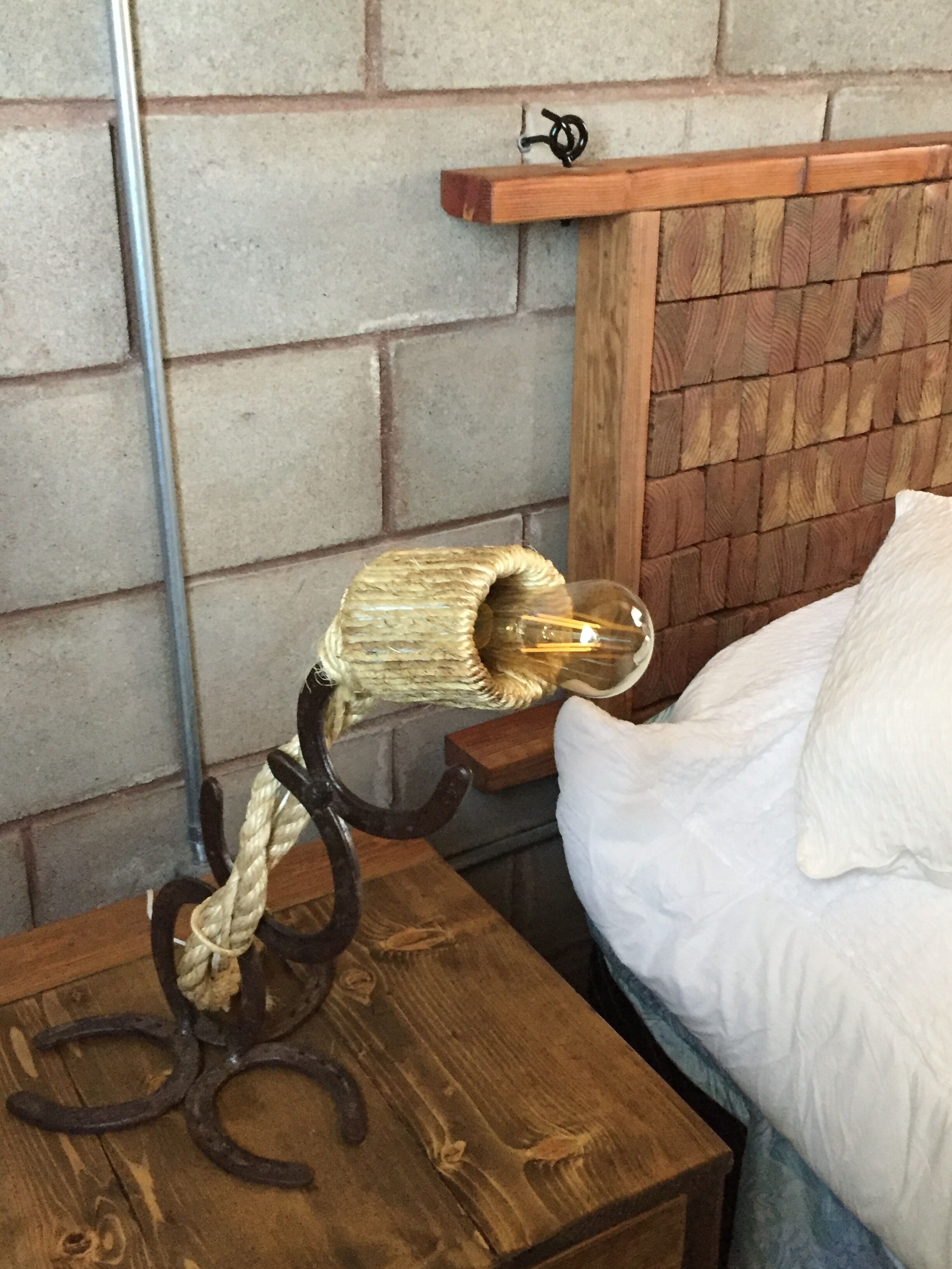 A wooden bedside table with a lamp featuring a rope and metal base, a brick wall, and a wooden headboard with a shelf.