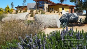 A modern house with large rock formations in the foreground, a clear blue sky, and a well-maintained garden with purple flowers. - Sn Tony Hotel & Spa (Valle de Guadalupe)