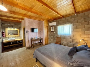 A cozy bedroom with a wooden ceiling, a bed with pillows, a bedside table, a mirror, a TV, and a brick wall. - Sn Tony Hotel & Spa (Valle de Guadalupe)