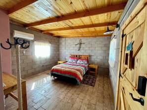 A bedroom with a wooden ceiling, a brick wall, a bed with a colorful quilt, a cat tree, and a wooden door with a handle.