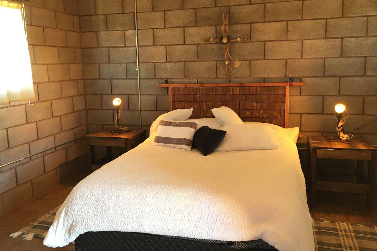A bedroom with a brick wall, a wooden headboard, a bed with white bedding, two bedside tables with lamps, and a window with curtains.