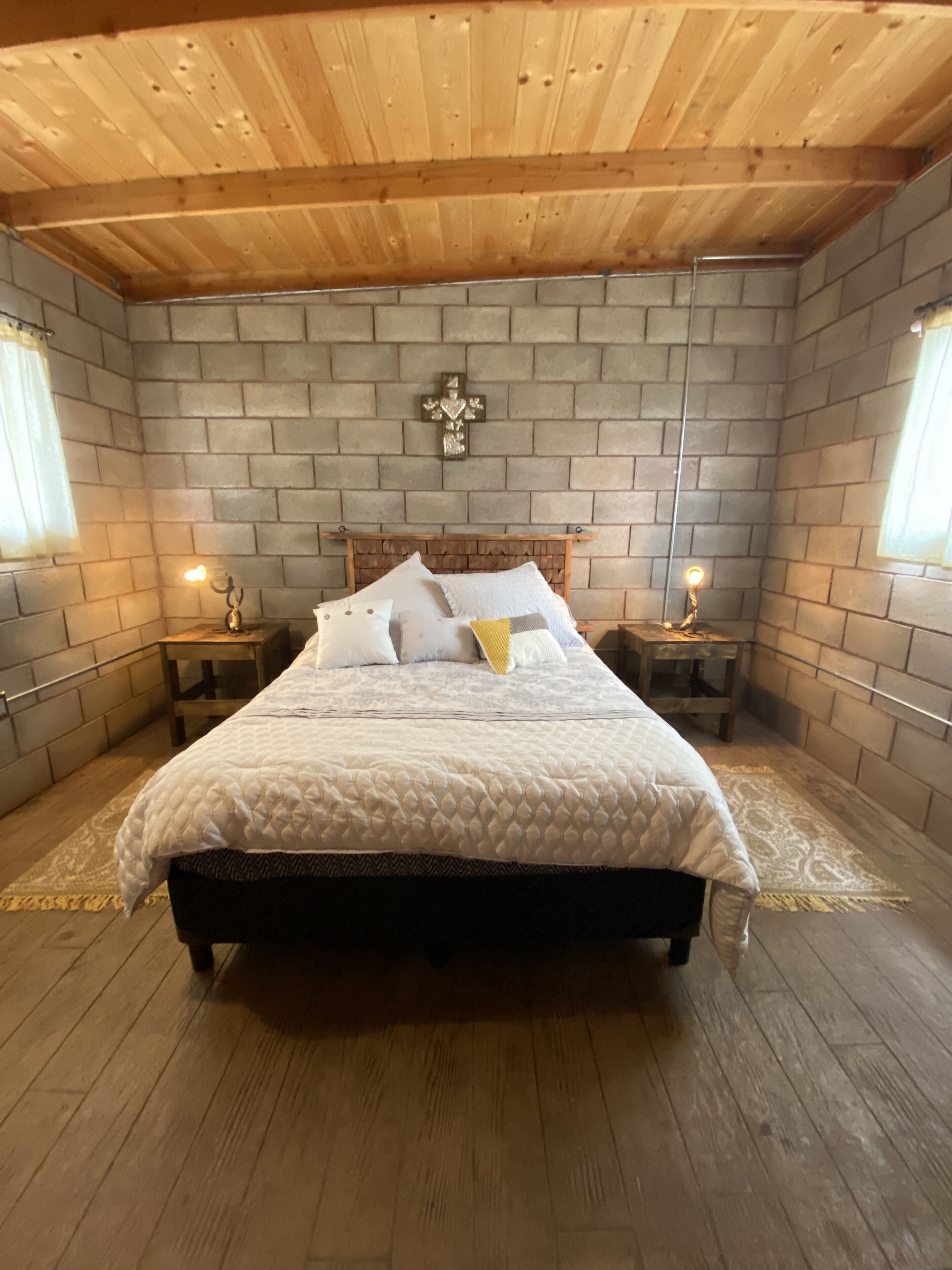 Un dormitorio con techo de madera, paredes de ladrillo, una decoración con cruz, una cama con ropa blanca, dos mesitas de noche con lámparas y una alfombra estampada.