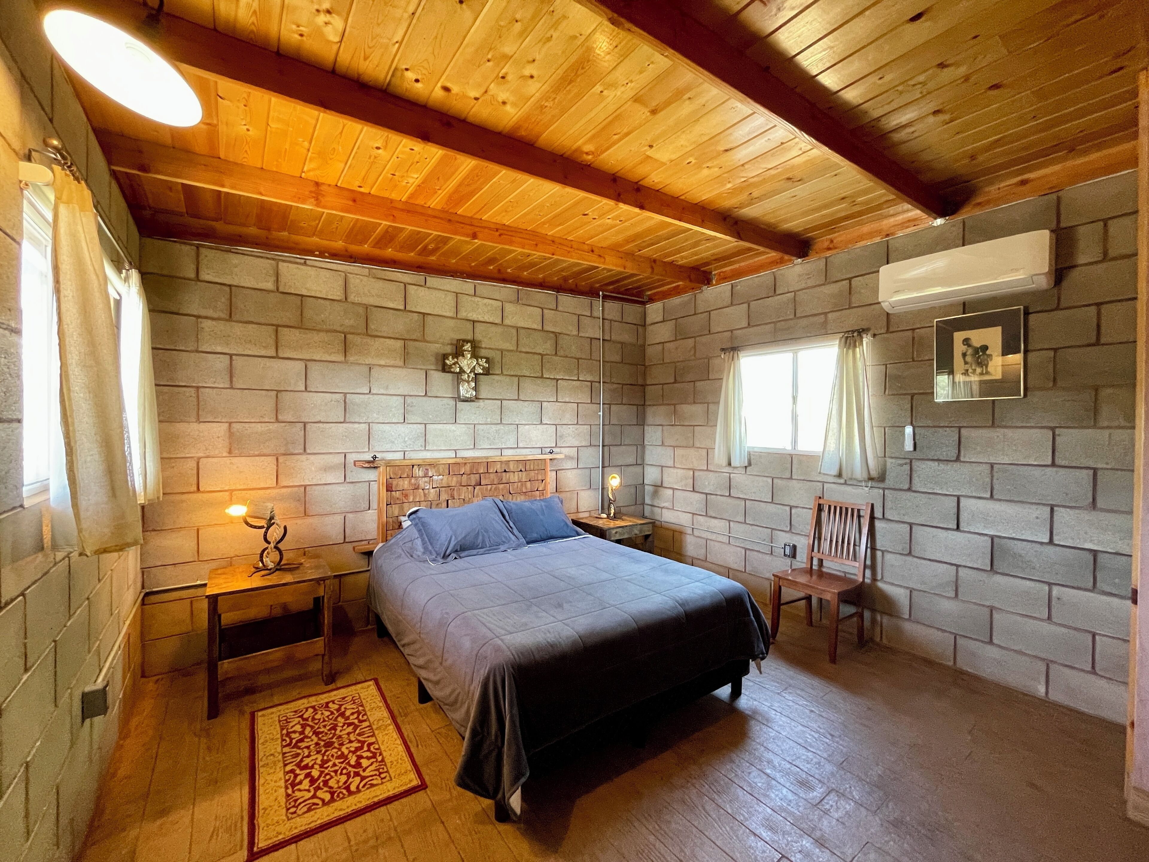 A bedroom with a wooden ceiling, brick walls, a bed with a headboard, a nightstand, a chair, a rug, and a picture on the wall.