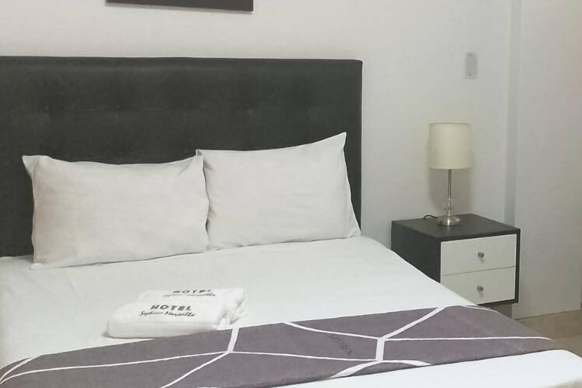 Ein ordentlich bezogenes Bett mit weißer Bettwäsche und einer Tagesdecke mit geometrischem Muster. Ein kleiner Nachttisch mit Lampe und Schublade steht daneben. Über dem Bett hängt ein gerahmtes Bild.