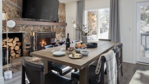 Un área de comedor con chimenea de piedra, un televisor de pantalla plana montado encima y un gran ventanal con cortinas.