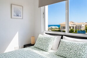 A bedroom with a bed, a window with a view of buildings and the sea, and a framed picture on the wall. - Narcissos Villa 2 - Three Bedroom Villa, Sleeps 8 (Protaras)