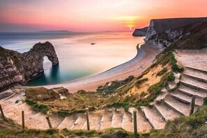 Un paisaje costero con una formación rocosa en arco natural, una playa y una serie de escalones que bajan hacia el agua.