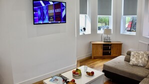 A modern living room with a flat-screen TV, a wooden cabinet, a sofa, and a coffee table with fruits and magazines. - Holiday home hot tub and sauna (Bournemouth)