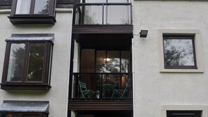 Un edificio de varios pisos con balcones, una ventana con vistas a árboles y un pequeño espacio al aire libre con sillas.
