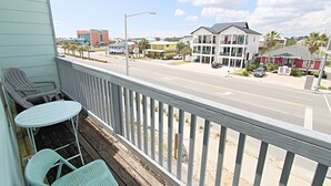 A balcony with a view of a street, a white building with a balcony, and a red building in the background. - 👣 Come get your toes in the sand ~~~ and a cold drink in your hand! 🍹 (Gulf Shores)