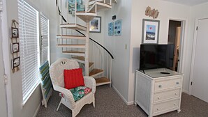 A room with a spiral staircase, a wicker chair with a red pillow, a dresser with a TV, and wall art. - 👣 Come get your toes in the sand ~~~ and a cold drink in your hand! 🍹 (Gulf Shores)