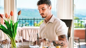 A man dining at an outdoor table with a view of the sea, a variety of food, and a vase of tulips.