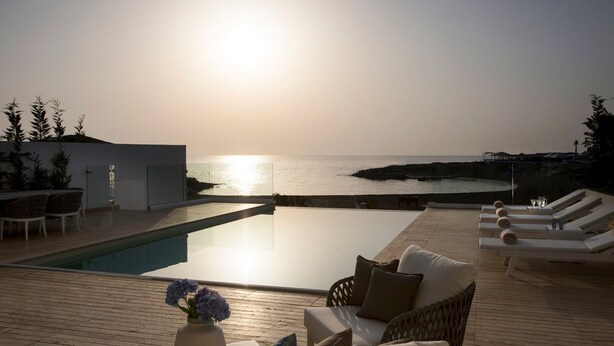 Un área junto a la piscina con sillones, una mesita con un florero y vistas al mar al atardecer.