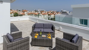 Una terraza en la azotea con mobiliario de mimbre, una mesa con una botella y vasos, y vistas de la ciudad.