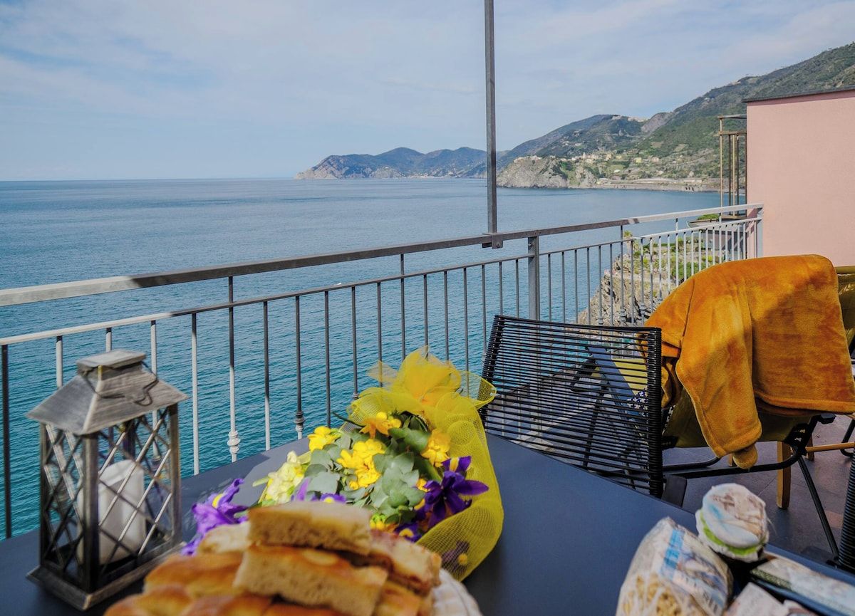 Un balcon avec vue sur la mer, une table garnie de nourriture et de fleurs, et une chaise avec une couverture.