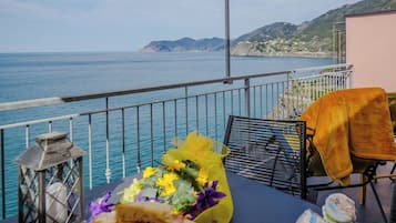 En balkong med utsikt över havet, ett bord med mat och blommor och en stol med en filt.