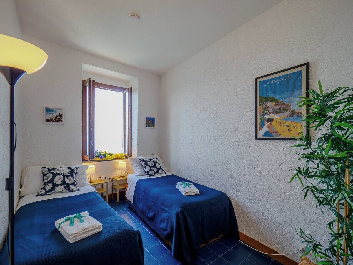 Una habitación con dos camas, un escritorio, una lámpara, un ventanal y un cuadro colgado en la pared.