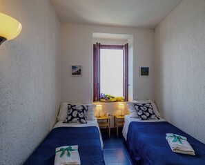 Ein kleiner Raum mit zwei Betten, einem Fenster mit Vorhängen, einem Schreibtisch mit Lampe und einem kleinen Tisch mit Blumenvase.