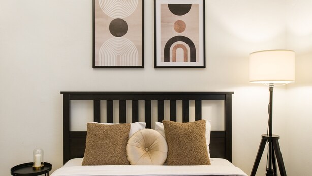 Una cama bien hecha con cabecera negra, dos cuadros abstractos enmarcados encima, una lámpara de noche y una mesita con una vela.