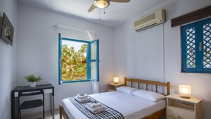 A bedroom with a bed, bedside tables, a desk, a chair, a window with an open door, and an air conditioning unit. - Protaras Ayios Elias Suite (Protaras)