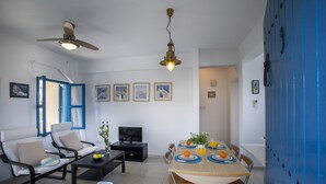A modern living room with a sofa, chairs, a dining table set with plates and glasses, a TV, and ceiling fans. - Protaras Ayios Elias Suite (Protaras)
