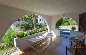 A sunlit patio with a hammock, a stone bench, and a view of a residential area through an archway.