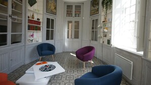 A room with a curved layout, featuring a white table with a book and a decorative plate, two blue chairs, a purple chair, and a bookshelf with various items and paintings. - 10' from St Rémy and Avignon, charming apartment ***, 2 bedrooms for 4 people (Noves)