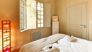 A hotel room with a bed, a radiator, a window with open shutters, and a bedside lamp. - 10' from St Rémy and Avignon, charming apartment ***, 2 bedrooms for 4 people (Noves)