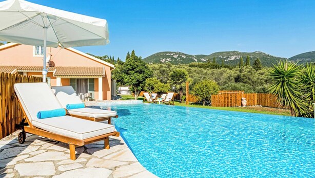A swimming pool with lounge chairs and a canopy, surrounded by a wooden fence and greenery, with mountains in the background. - Villa Secret Paradise (Corfu)