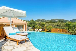 Piscina con sillas de descanso y una pérgola, rodeada de una valla de madera y follaje, con montañas al fondo.