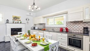 A modern kitchen with white cabinetry, a dining table set with breakfast, and a fireplace. - Villa Secret Paradise (Corfu)