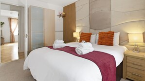 Quarto de hotel com uma cama grande, cabeceira de madeira, mesa de cabeceira com abajur e porta que dá para outro aposento.