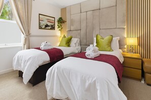 Une chambre d’hôtel avec deux lits, une grande tête de lit rembourrée, une table de chevet avec une lampe et une fenêtre avec des rideaux.