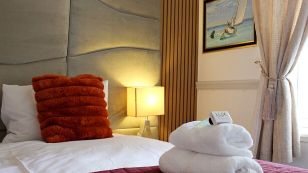 Quarto de hotel com uma cama, almofadas, candeeiro, quadro e janela com cortinas.