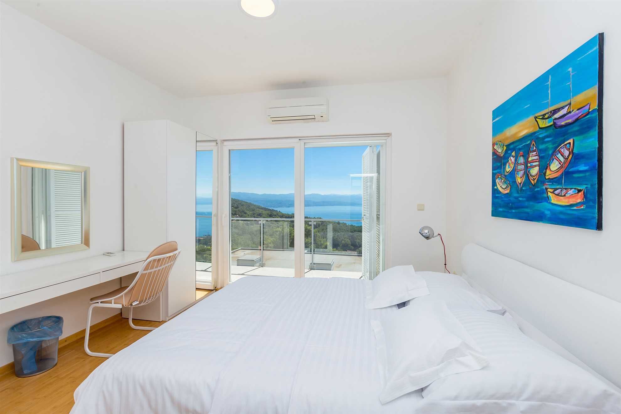 A bedroom with a large bed, a desk with a chair, a window with a view of the sea, and a painting on the wall.