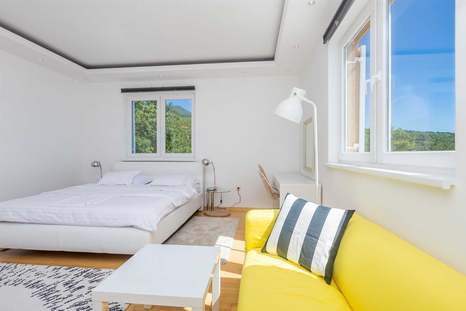 A bedroom with a bed, a yellow sofa, a desk, and a window with a view of greenery.