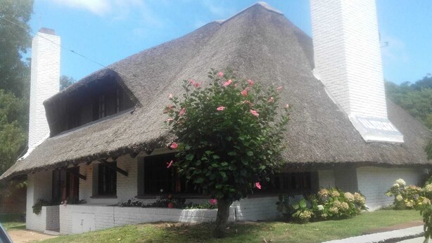Una casa tradizionale con tetto di paglia, un camino, un portico con una panchina e un albero con fiori rosa di fronte.