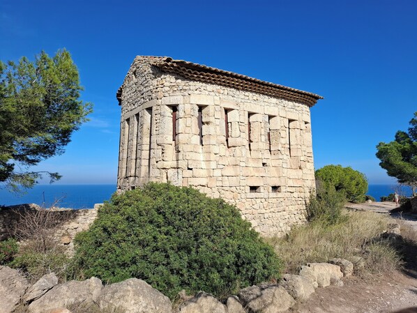A stone structure with a tiled roof, surrounded by greenery and overlooking a body of water.